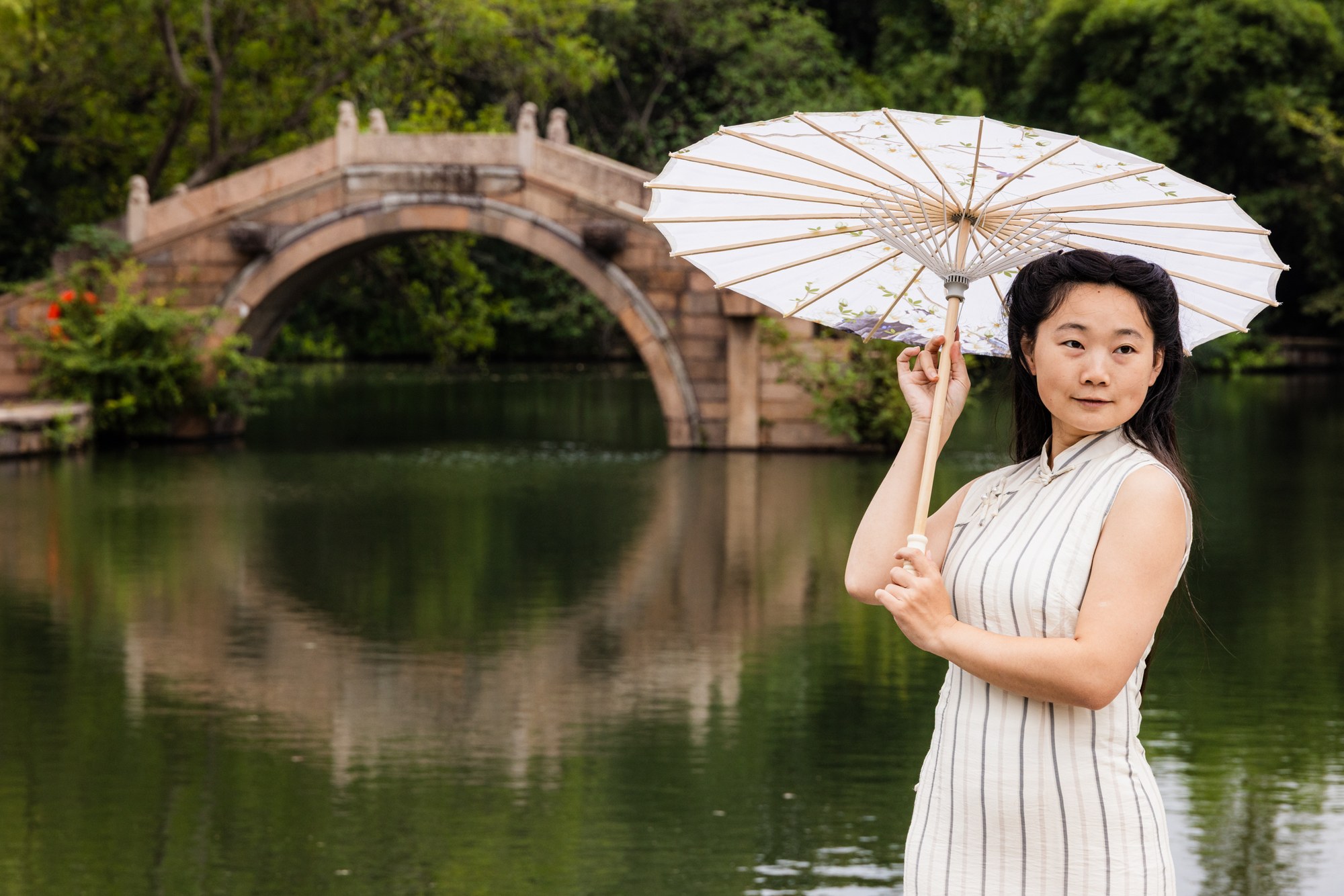 Woman in a qipao with a white parasol by the moon bridge, reflected in the water, China