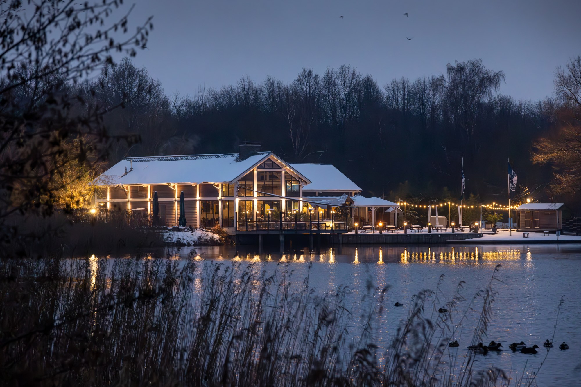 Lit pavilion by a snow-covered lake in the blue hour, warm lights reflected in the water, Netherlands