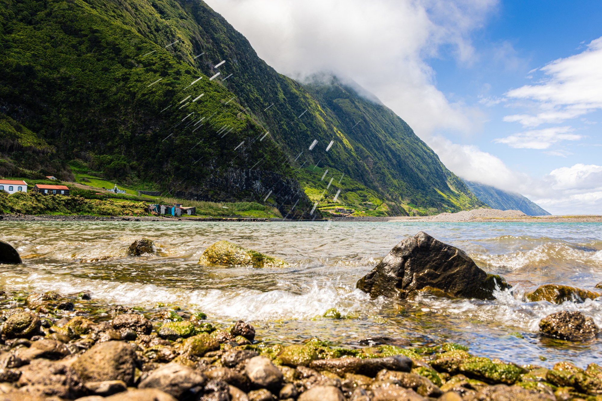 Waves breaking over mossy rocks on the fajã coast, ocean spray in the air, green cliff face and turquoise bay, Azores
