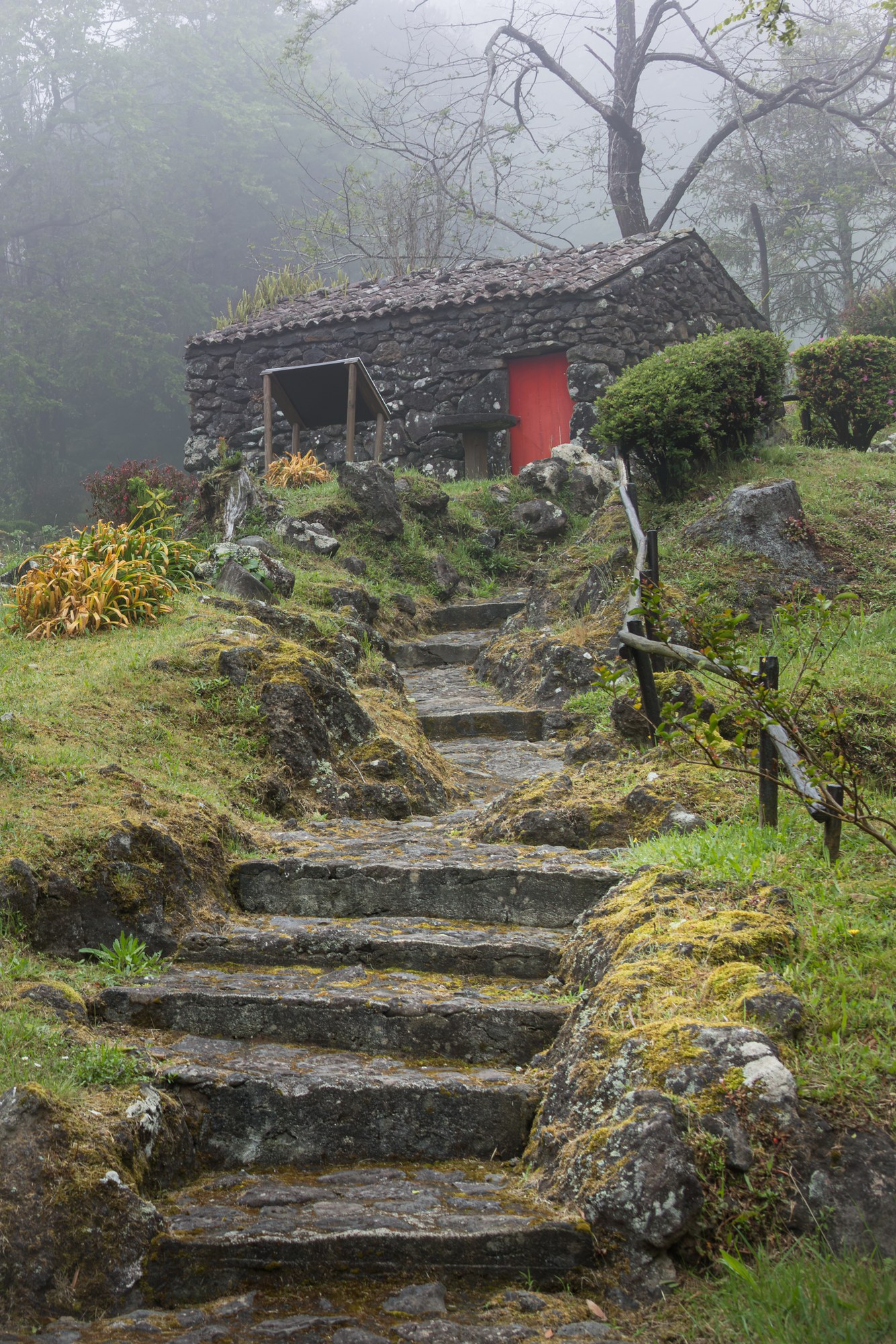 A mossy natural-stone stairway leading up to a stone hut with a red door in the mist, Azores