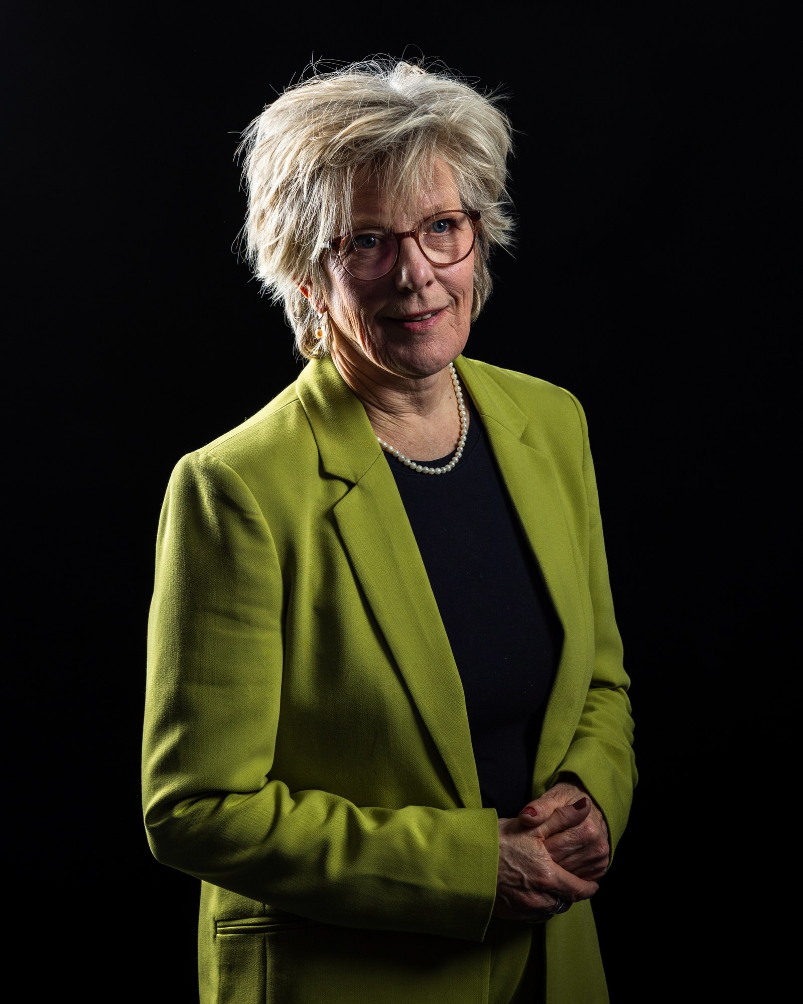 Older woman in a chartreuse blazer with a pearl necklace, gentle smile, studio portrait on black