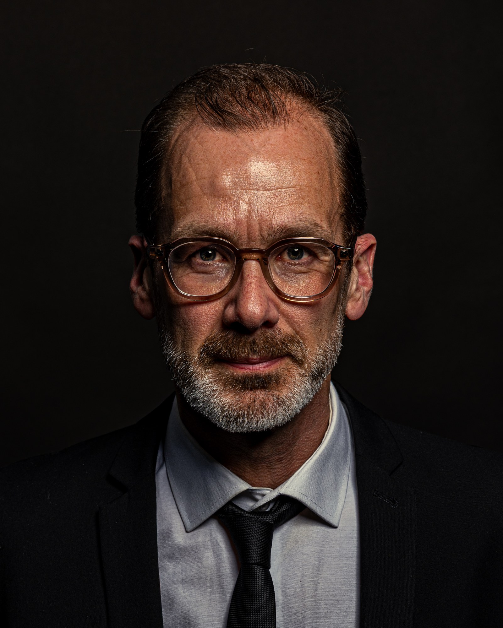 Man with clear-framed glasses looking straight into the lens, studio portrait on black